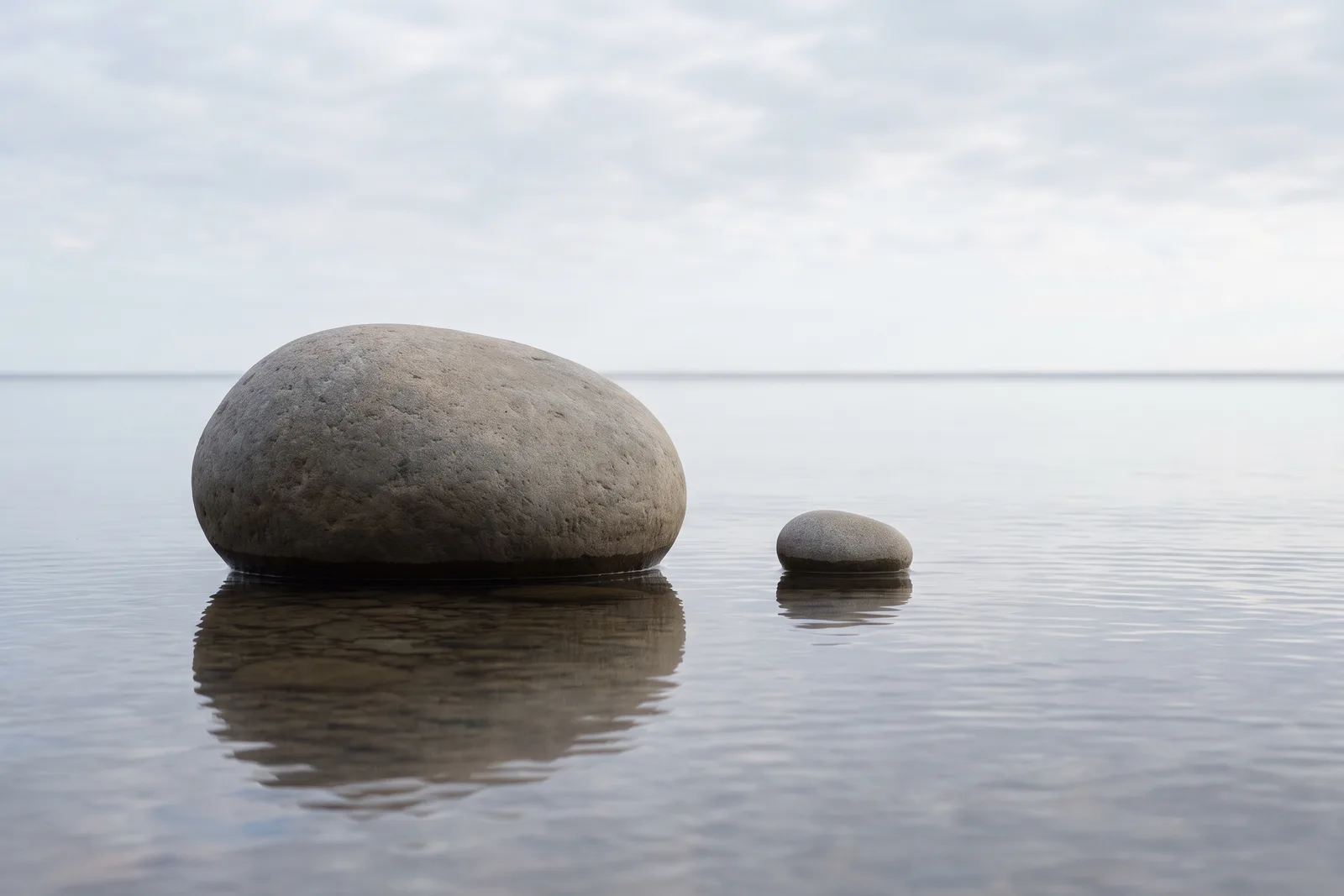 Two identically shaped stones side by side in shallow mirror-calm water — one enormous, one tiny — both perfectly reflected.